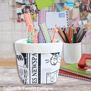 Puede incluir: Un contenedor de almacenamiento de lona blanca con estampado de periódico en blanco y negro. El contenedor está lleno de útiles escolares coloridos, incluyendo reglas, tijeras, lápices y un bolígrafo rosa de flamenco.