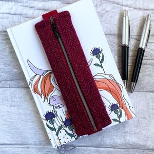 May include: A burgundy pencil case with a silver zip lies on a white notebook with a Highland cow and thistle design. Two black and silver pens are also visible. The pencil case is made of a textured fabric.
