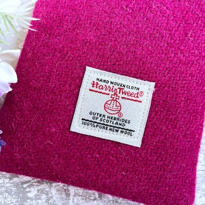 Pu&ograve; includere: Una pochette in tessuto Harris Tweed color magenta vivace con un'etichetta rettangolare bianca tessuta. L'etichetta riporta il testo HAND WOVEN CLOTH, Harris Tweed, OUTER HEBRIDES OF SCOTLAND e 100% PURE NEW WOOL, con un emblema circolare.