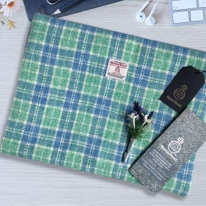 May include: A blue and green plaid throw blanket made of Harris Tweed wool. The blanket is folded and displayed on a white wooden surface. The label on the blanket reads "Harris Tweed Handwoven in the Outer Hebrides of Scotland from Pure Wool."