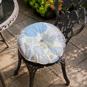 May include: A round, white seat cushion featuring a light blue and olive green floral pattern with a central button, placed on a black metal chair with a rose-motif backrest. The cushion is tufted and fits the circular seat of the chair.
