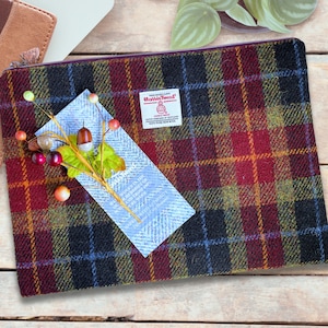May include: A rectangular zippered pouch made of Harris Tweed fabric featuring a dark plaid pattern in burgundy, black, and yellow. A white label with the Harris Tweed logo is sewn on the front, and a decorative sprig of artificial berries is attached.