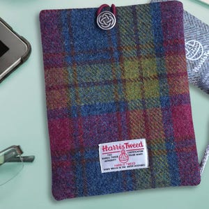 May include: A Harris Tweed tablet case in a plaid design of blue, purple, and green. The case has a silver-coloured button with a Celtic knot design. A white label with red text reads "Harris Tweed". The case is designed to hold a tablet.