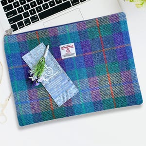 May include: A rectangular Harris Tweed zip pouch featuring a blue, purple, and green plaid pattern. A white label with the Harris Tweed logo and text is attached to the front, alongside a small decorative sprig of dried flowers tied with twine.