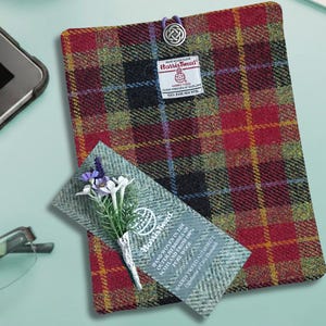 Op de afbeelding: Een Harris Tweed tablethoes in een rood, zwart en geel geruit patroon. Een klein label met het Harris Tweed-logo is bevestigd. Een klein takje bloemen is op een bijpassende stofstaal geplaatst.