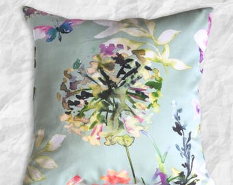 Könnte beinhalten: Ein quadratisches Zierkissen mit hellsalbeigrünem Hintergrund und einem Aquarell-Druck von Blumen und Schmetterlingen in Rosa, Violett, Gelb und Dunkelgrün. Der Text Cotton & Tweed Designs ist unten in violetter Farbe aufgedruckt.