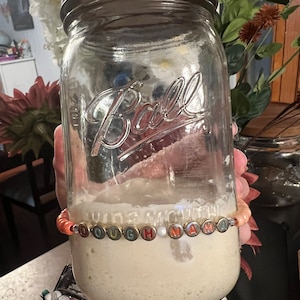 Pode incluir: Um frasco de vidro transparente com tampa prateada, preenchido com uma mistura de cor clara. Uma pulseira de contas com a inscrição "DOUGH MAMA" circunda o frasco. O frasco é segurado em frente a um fundo desfocado.