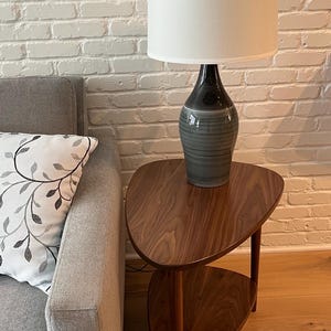 May include: A table lamp with a white drum shade and a teardrop-shaped ceramic base featuring a gradient from black at the top to a grey and white horizontal striped pattern at the bottom, resting on a dark wood side table with a lower shelf.