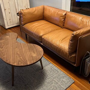 May include: A brown leather sofa with a wooden frame and a unique kidney-shaped wooden coffee table with a dark wood finish. The sofa is sitting on a gray rug with a geometric pattern.