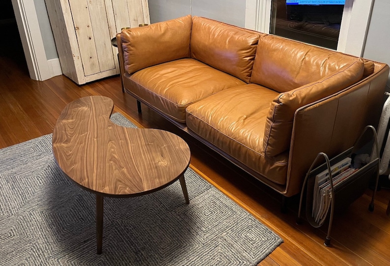 May include: A brown leather sofa with a wooden frame and a unique kidney-shaped wooden coffee table with a dark wood finish. The sofa is sitting on a gray rug with a geometric pattern.