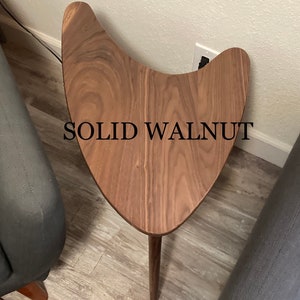 May include: A solid walnut wood side table with a unique boomerang shape and a single leg. The table is shown in a home setting.