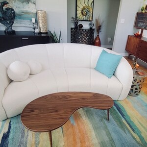 May include: A white curved sofa with two white pillows and a light blue throw pillow. The sofa is in a living room with a wooden coffee table and a colorful rug.