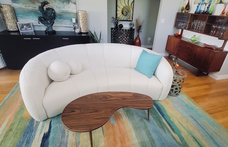 May include: A white curved sofa with two white pillows and a light blue throw pillow. The sofa is in a living room with a wooden coffee table and a colorful rug.