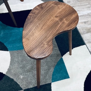 May include: A wooden kidney-shaped side table with a dark brown stain. The table has three legs and is sitting on a rug with a geometric pattern.