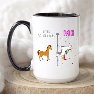 Puede incluir: Taza de café blanca con borde negro. La taza tiene un gráfico de un unicornio haciendo una danza de poste con el texto "ME" encima. El texto "OTHER 50 YEAR OLDS" está en el lado izquierdo de la taza con un gráfico de un caballo marrón.