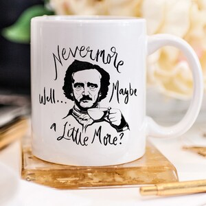 Puede incluir: Taza de cer&aacute;mica blanca con una ilustraci&oacute;n en blanco y negro de Edgar Allan Poe sosteniendo una taza de t&eacute;. La taza tiene el texto "Nevermore well... maybe a little more?" impreso en ella.