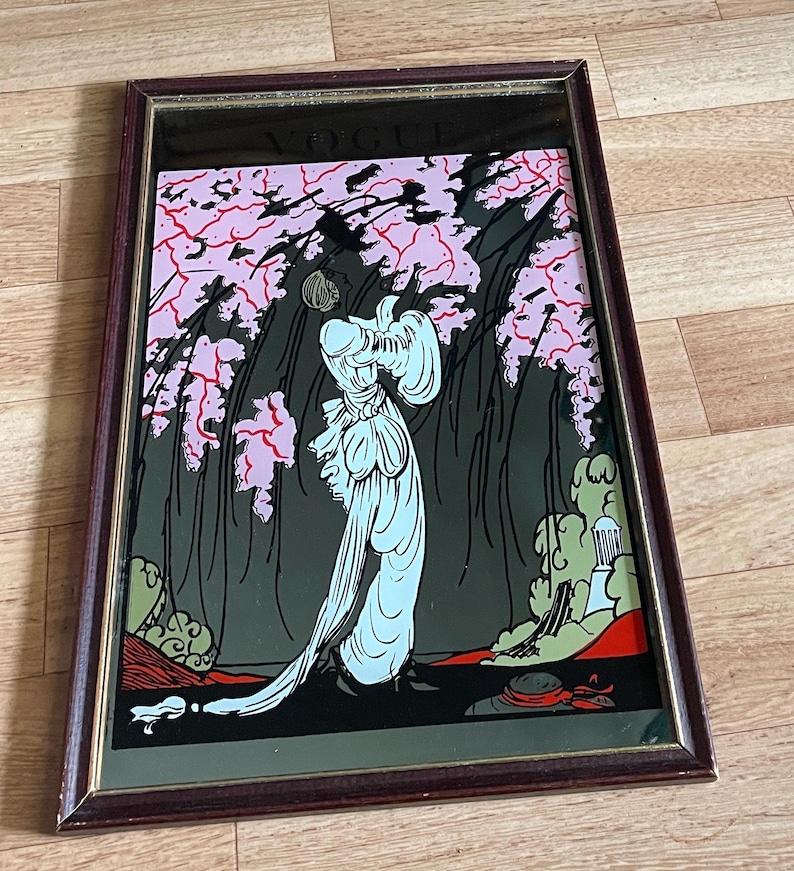May include: A framed art print featuring a woman in a flowing gown beneath pink flowering trees. The artwork is set against a dark background with green and red accents. The frame is a dark brown color.