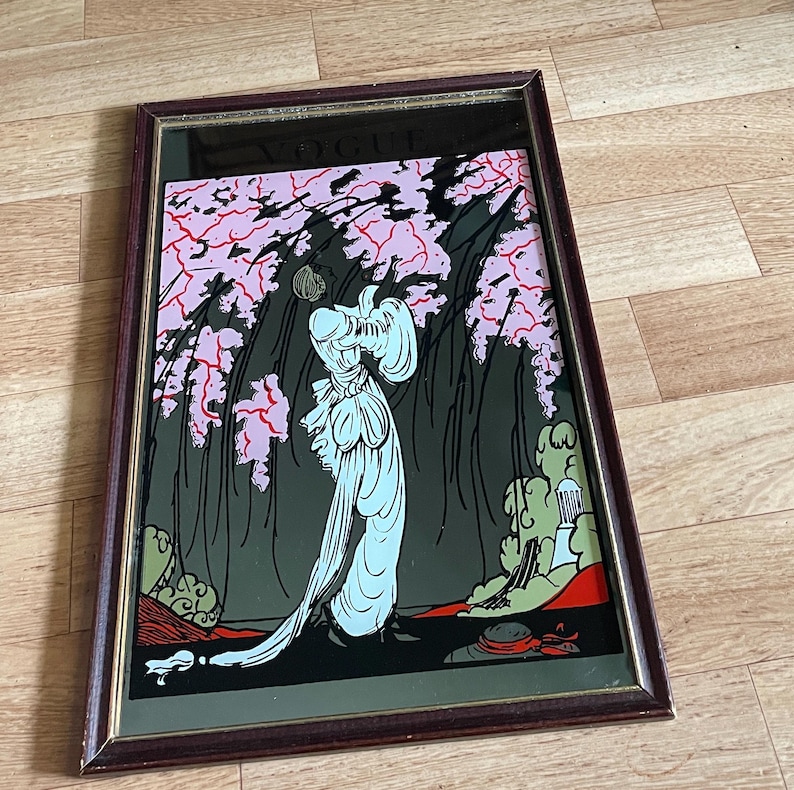 May include: A framed vintage art deco mirror featuring a stylized illustration of a woman in a flowing gown beneath pink flowering branches. The artwork is set against a dark background, with a brown frame. The word "VOGUE" is visible at the top.