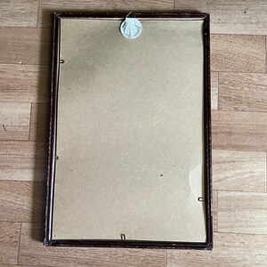 May include: A rectangular picture frame with a dark brown wooden frame. The frame holds a beige backing and is secured with small metal clips. A white adhesive sticker is attached to the back for hanging.