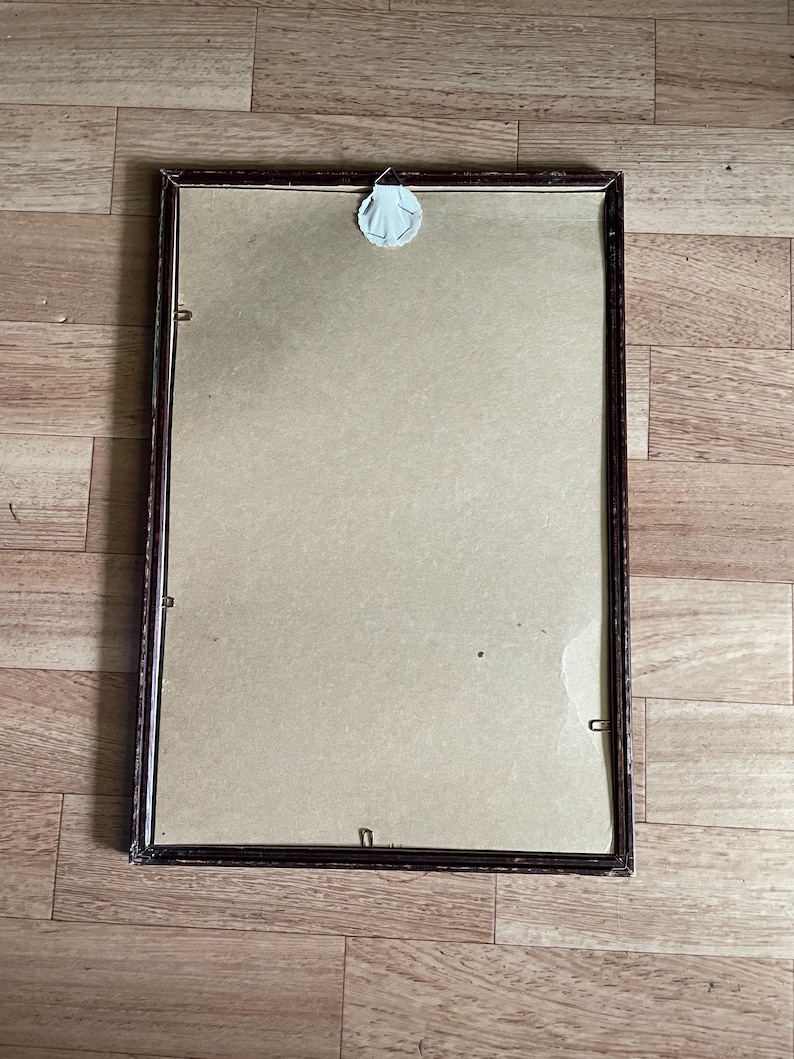 May include: A rectangular picture frame with a dark brown wooden frame. The frame holds a beige backing and is secured with small metal clips. A white adhesive sticker is attached to the back for hanging.