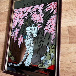 May include: Vintage framed mirror with a decorative art deco design. The mirror features the word "VOGUE" at the top. The artwork depicts a woman in a flowing white gown, set against a backdrop of pink blossoms and stylized foliage.