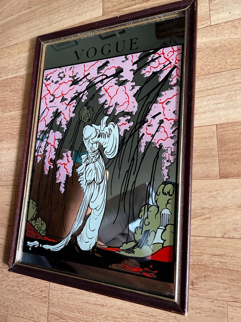 May include: Vintage framed mirror with a decorative art deco design. The mirror features the word "VOGUE" at the top. The artwork depicts a woman in a flowing white gown, set against a backdrop of pink blossoms and stylized foliage.