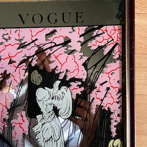 May include: A framed mirror with the word "VOGUE" at the top. The mirror features a pink floral design with black accents and a stylized illustration of a person in white. The frame is a dark brown color.