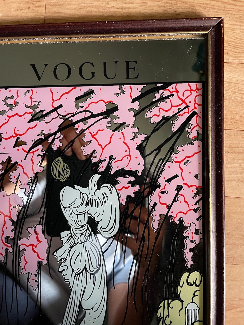 May include: A framed mirror with the word "VOGUE" at the top. The mirror features a pink floral design with black accents and a stylized illustration of a person in white. The frame is a dark brown color.