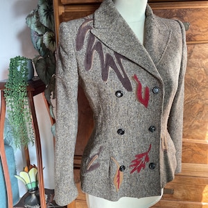 Op de afbeelding: Een bruine tweed blazer met een dubbele rij knopen en decoratieve bladvormige applicaties in tinten bruin, rood en beige.