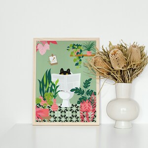 May include: A framed art print featuring a black cat reading a newspaper while sitting on a toilet, surrounded by lush green plants and pink flowers. The artwork has a light green background and is in a wooden frame. A white vase with dried flowers is to the right.