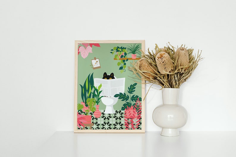 May include: A framed art print featuring a black cat reading a newspaper while sitting on a toilet, surrounded by lush green plants and pink flowers. The artwork has a light green background and is in a wooden frame. A white vase with dried flowers is to the right.