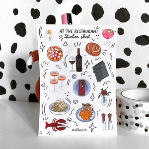 May include: A sticker sheet with a white background and black outlines. The sheet features illustrations of food, drinks, and other restaurant-themed items, including a glass of wine, a plate of pasta, a loaf of bread, a cocktail, and a menu. The text "At the Restaurant... Sticker Sheet" is written at the top of the sheet.
