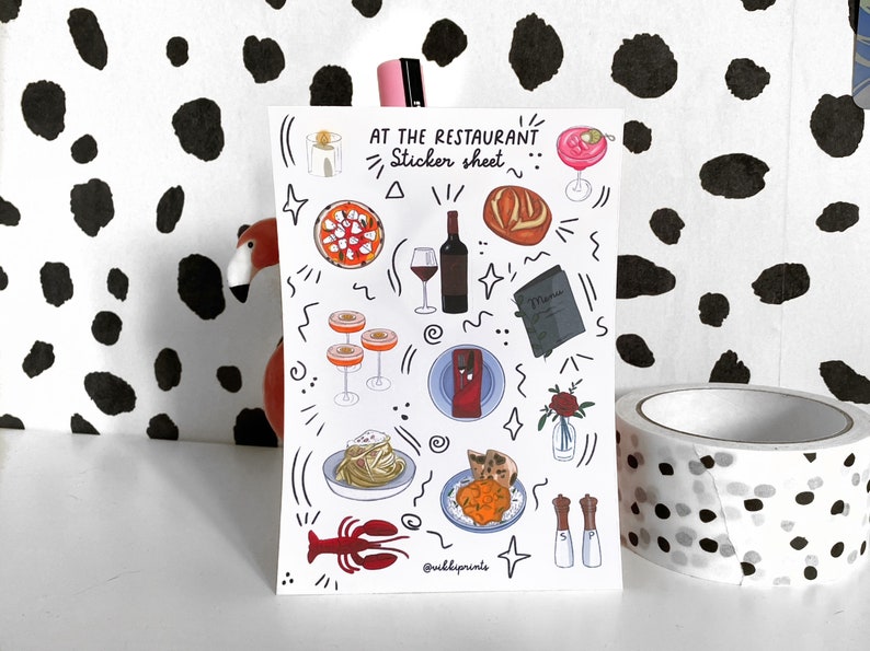 May include: A sticker sheet with a white background and black outlines. The sheet features illustrations of food, drinks, and other restaurant-themed items, including a glass of wine, a plate of pasta, a loaf of bread, a cocktail, and a menu. The text "At the Restaurant... Sticker Sheet" is written at the top of the sheet.