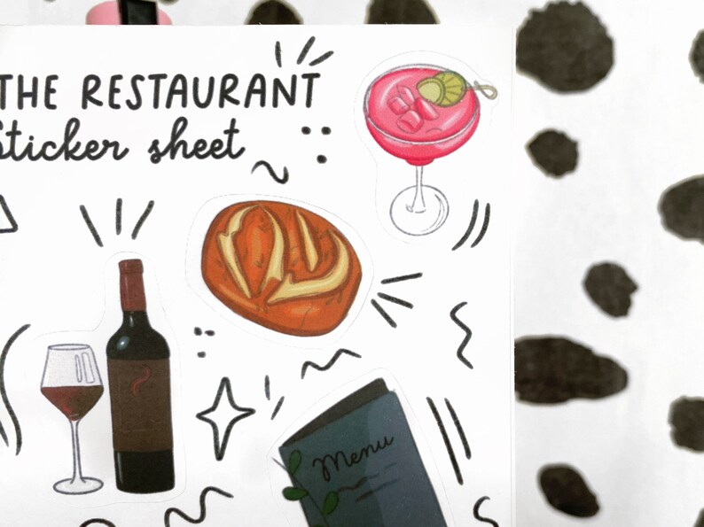 May include: A sticker sheet featuring a selection of restaurant-themed illustrations, including a loaf of bread, a glass of wine, a bottle of wine, a cocktail, and a menu. The sheet is white with black outlines and text that reads "The Restaurant Sticker Sheet".