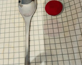 May include: A stainless steel spoon with a long handle and a rounded bowl. The spoon is on a white and gray checkered surface, with a red circular object, a yellow wavy shape, and a teal triangle.