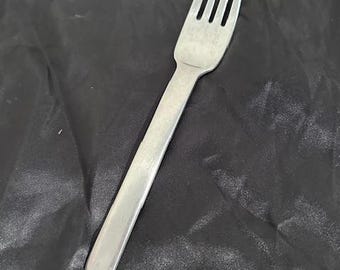 May include: A stainless steel fork with four tines and a long, rectangular handle. The fork is positioned diagonally against a dark, textured background, suggesting a dining or kitchen setting.