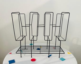 May include: A black metal magazine rack with a geometric design. The rack has four vertical compartments and a horizontal shelf at the bottom. The rack is made of thin, black metal rods and is placed on a table with a white and colorful patterned tablecloth.