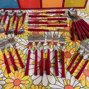 Puede incluir: Un juego de cubiertos vintage con mangos rojos, que incluye cucharas, tenedores y cuchillos. La cubertería está dispuesta sobre un mantel con estampado floral con flores amarillas, naranjas y blancas en forma de margarita.