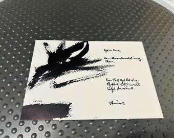 May include: A rectangular white card featuring a bold black abstract ink brushstroke on the left side. The text reads: You are an emanating star in the galaxy of the eternal life divine. The word Corita is printed near a black bar at the bottom.