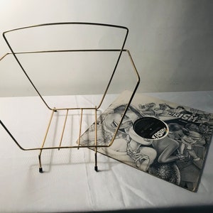 Puede incluir: Un estante de discos de metal dorado con un disco de vinilo negro apoyado en él. La portada del disco presenta una ilustración en blanco y negro de una banda con la palabra "PRISM" impresa en ella.