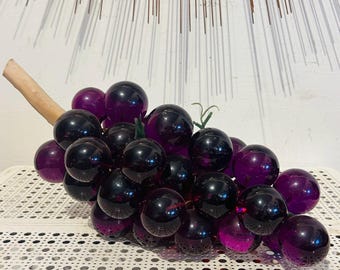 May include: A decorative cluster of translucent, deep purple glass grapes attached to a light-colored wooden stem. The individual spherical grapes vary slightly in size and depth of color, creating a realistic representation of a bunch of fruit.