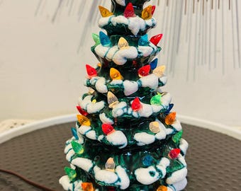 May include: A ceramic tabletop Christmas tree with a glossy dark green finish and textured white snow accents on the branches. The tree is decorated with numerous small, multi-colored plastic light bulbs in red, blue, yellow, and clear tones.