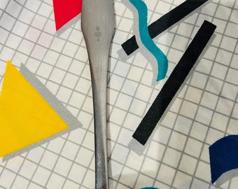 May include: A stainless steel butter knife with a long handle. The knife is on a white patterned surface with geometric shapes in red, yellow, black, and blue. The knife has a slightly worn appearance.