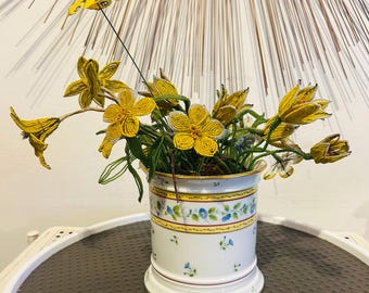 May include: A white ceramic cylindrical planter with a floral pattern of blue flowers and green leaves, featuring a yellow band near the top rim. The planter holds a bouquet of yellow beaded flowers and a yellow beaded butterfly on a wire stem.