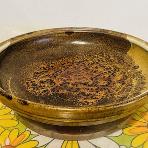 May include: A round, ceramic bowl with a brown and gold speckled design. The bowl has a wide rim and a textured interior. The bowl is sitting on a wooden base and a floral patterned tablecloth.