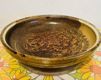 May include: A round, ceramic bowl with a brown and gold speckled design. The bowl has a wide rim and a textured interior. The bowl is sitting on a wooden base and a floral patterned tablecloth.