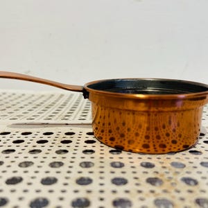 May include: A small, round copper-coloured saucepan with a single long, flat handle extending from the side. The exterior has a metallic, reflective finish, while the interior is dark and circular, featuring a smooth, uniform rim around the top edge.