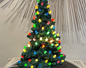 May include: A dark green ceramic Christmas tree with a textured, molded surface. The tree is illuminated by numerous small, multi-colored plastic bulbs in shades of red, blue, yellow, and pink, with a single clear bulb at the very top point.