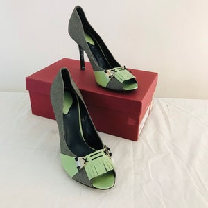 May include: A pair of green and black peep-toe heels with a fringe detail and a gold buckle. The shoes are on a red box.