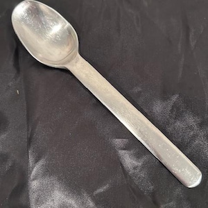 May include: A silver spoon with a long handle and an oval bowl. The spoon is made of a shiny metal and is set against a dark background. The spoon is likely for eating or serving food.
